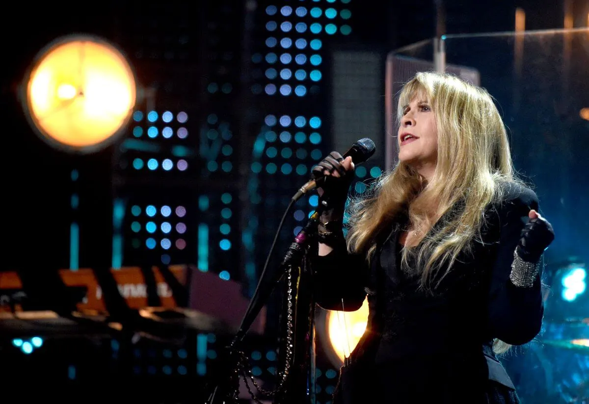 Stevie Nicks stands on a stage and holds a microphone. She wears a black dress and black fingerless gloves.