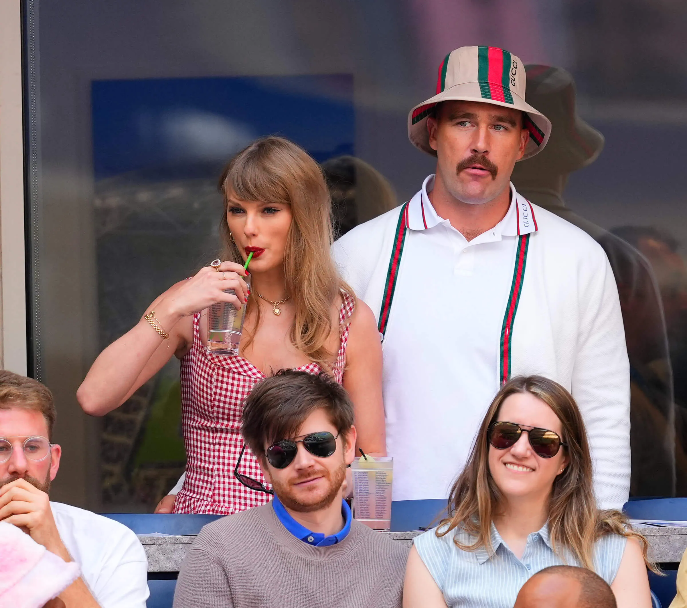 Taylor Swift taking a drink through a straw while next to Travis Kelce at the US Open. Swift is wearing a red plaid dress. Kelce is wearing a white button-up shirt with striped suspenders and a matching hat. Others sit in front of them.