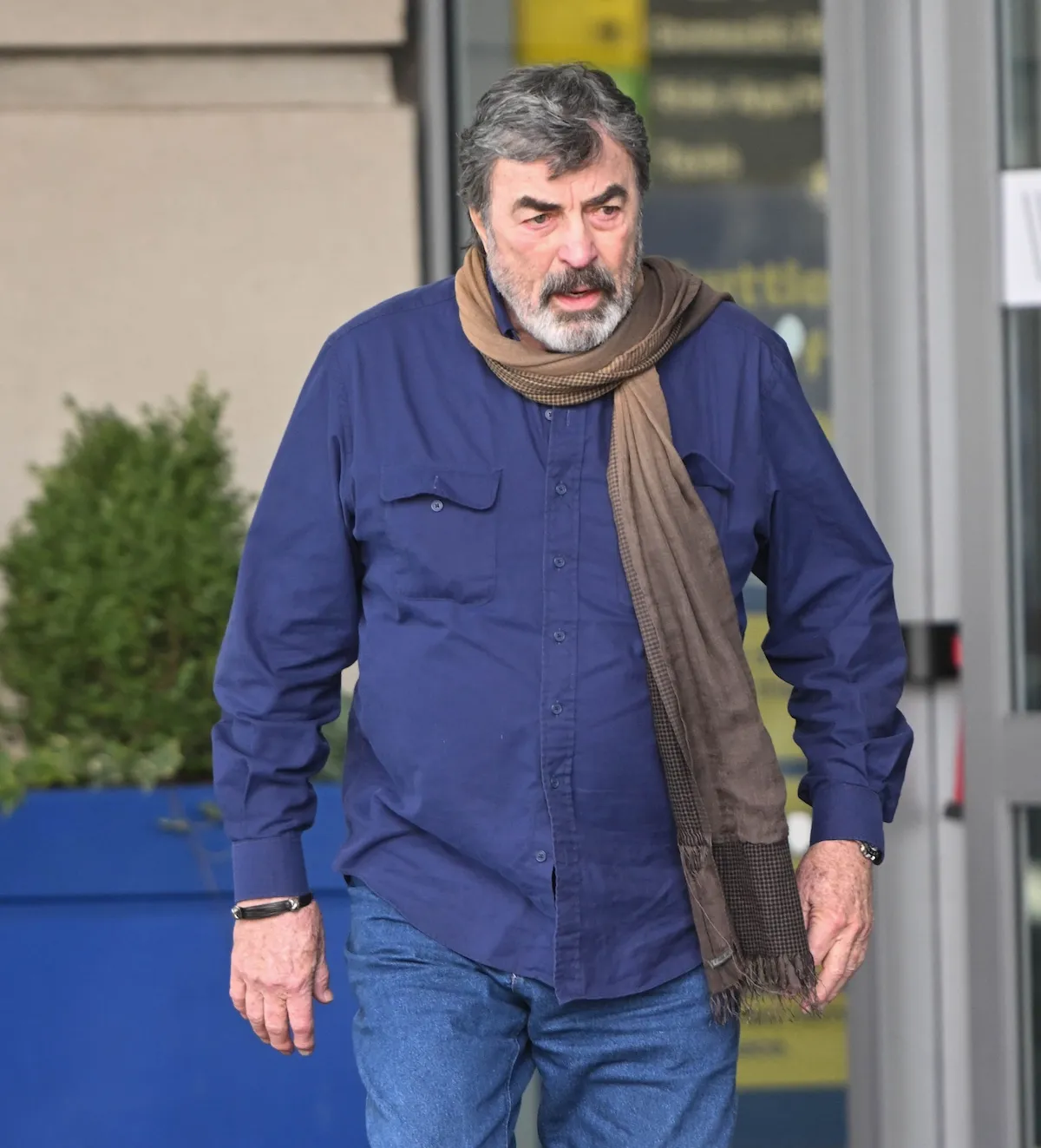 Tom Selleck, wearing a blue button-down shirt and a scarf, outside of JFK airport