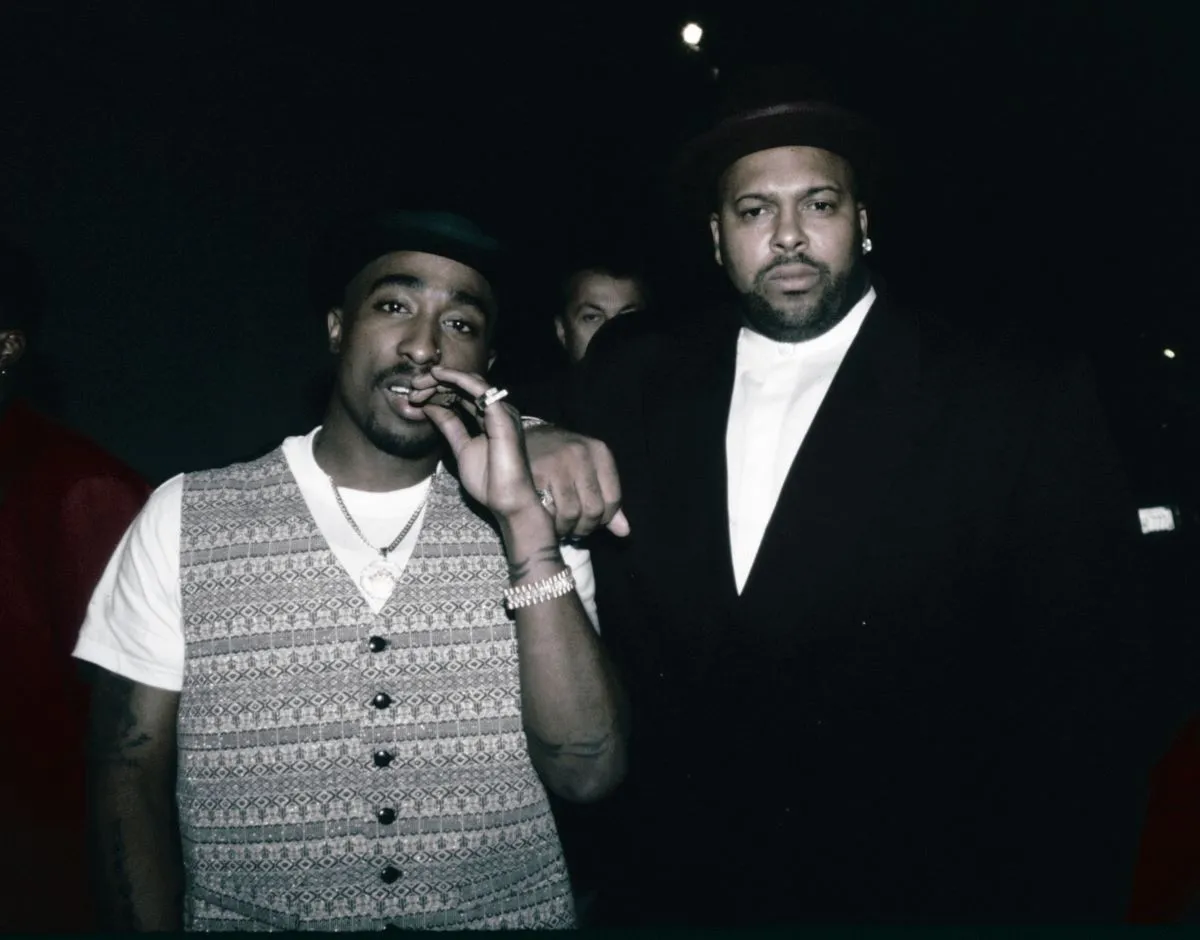 Tupac Shakur and Suge Knight stand together. Suge Knight has his arm on Tupac's shoulder.