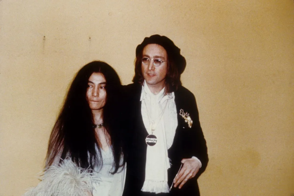 Yoko Ono and John Lennon stand against a yellow wall. Lennon wears a suit and sunglasses. Ono wears a white dress.