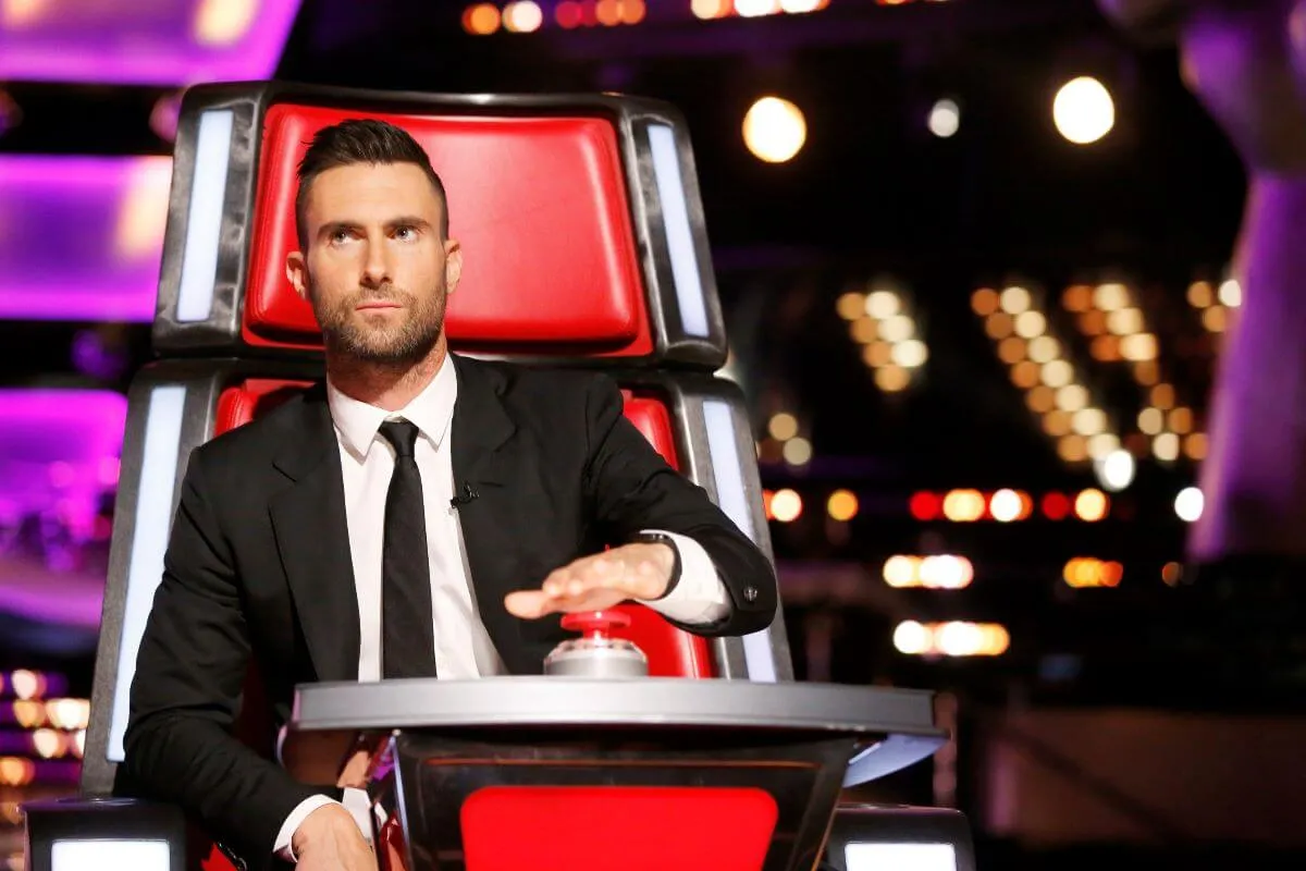 Adam Levine wears a suit and tie and sits in a red chair on 'The Voice.'