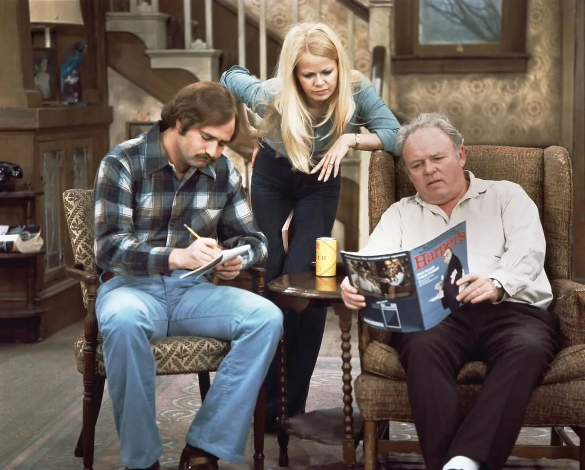 Archie Bunker reading an issue of Harper's with Gloria looking over his shoulder and Mike sitting next to him in 'All In The Family'