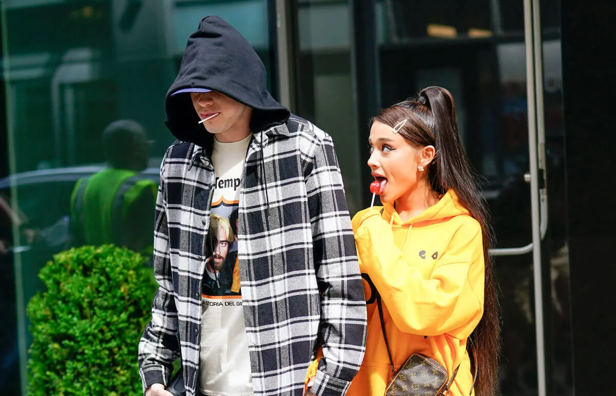 Pete Davidson wears a plaid shirt and hoodie while walking with Ariana Grande, who wears a yellow sweatshirt. They both have lollipops.