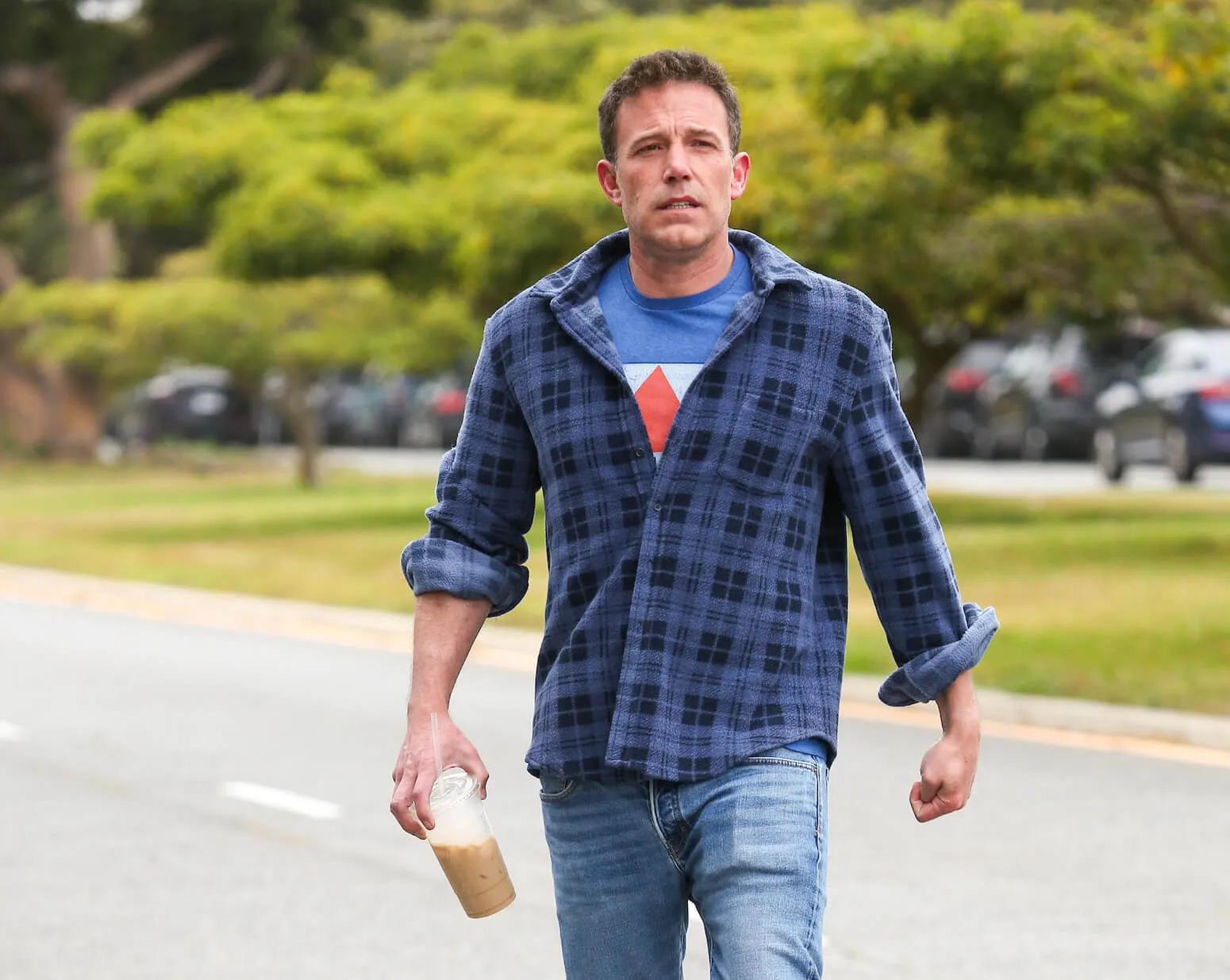 Ben Affleck walking on the street in Los Angeles. He's wearing a plaid shirt and jeans and holding an iced coffee.