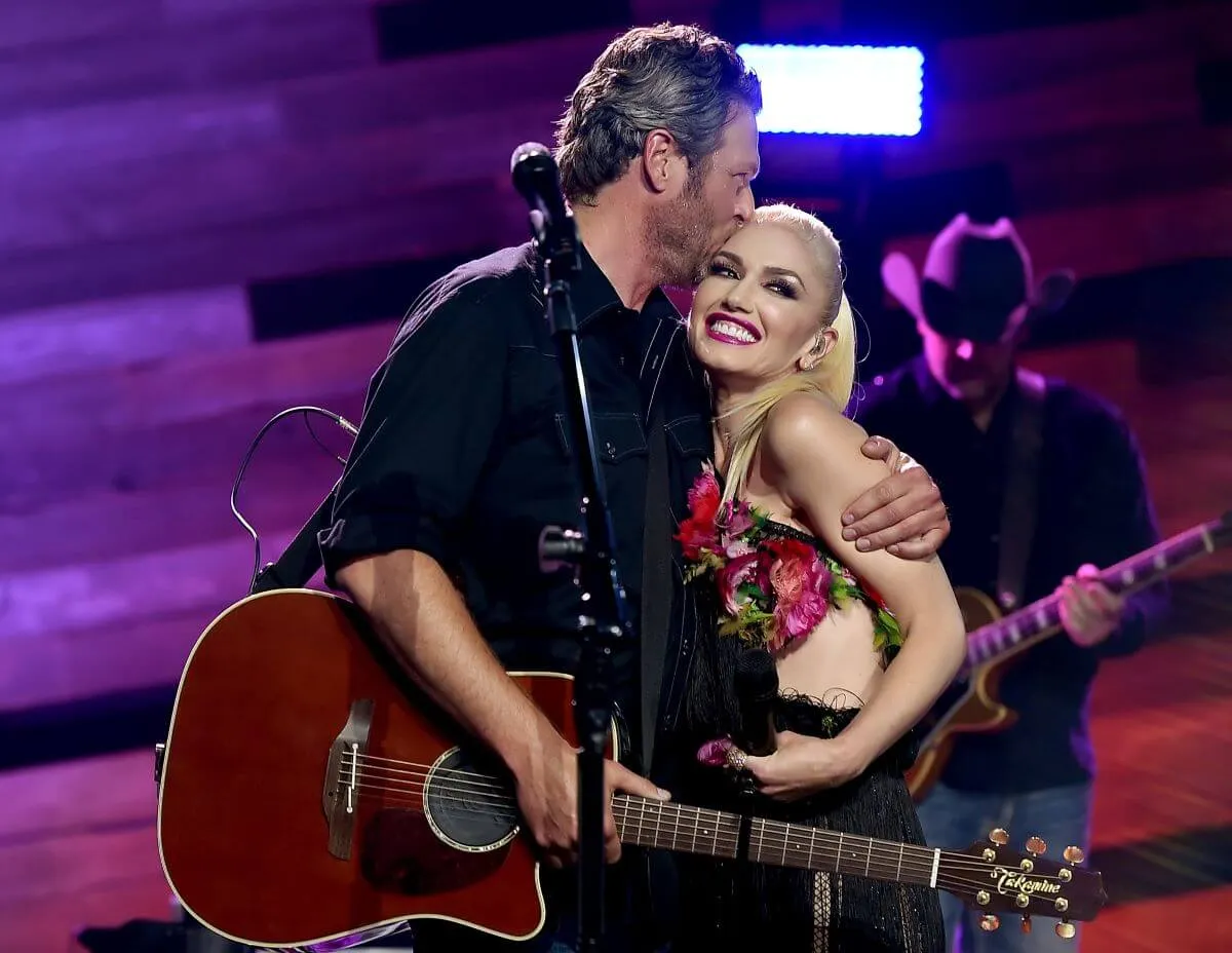 Blake Shelton holds a guitar and wraps his arm around Gwen Stefani while on stage. He kisses her head.