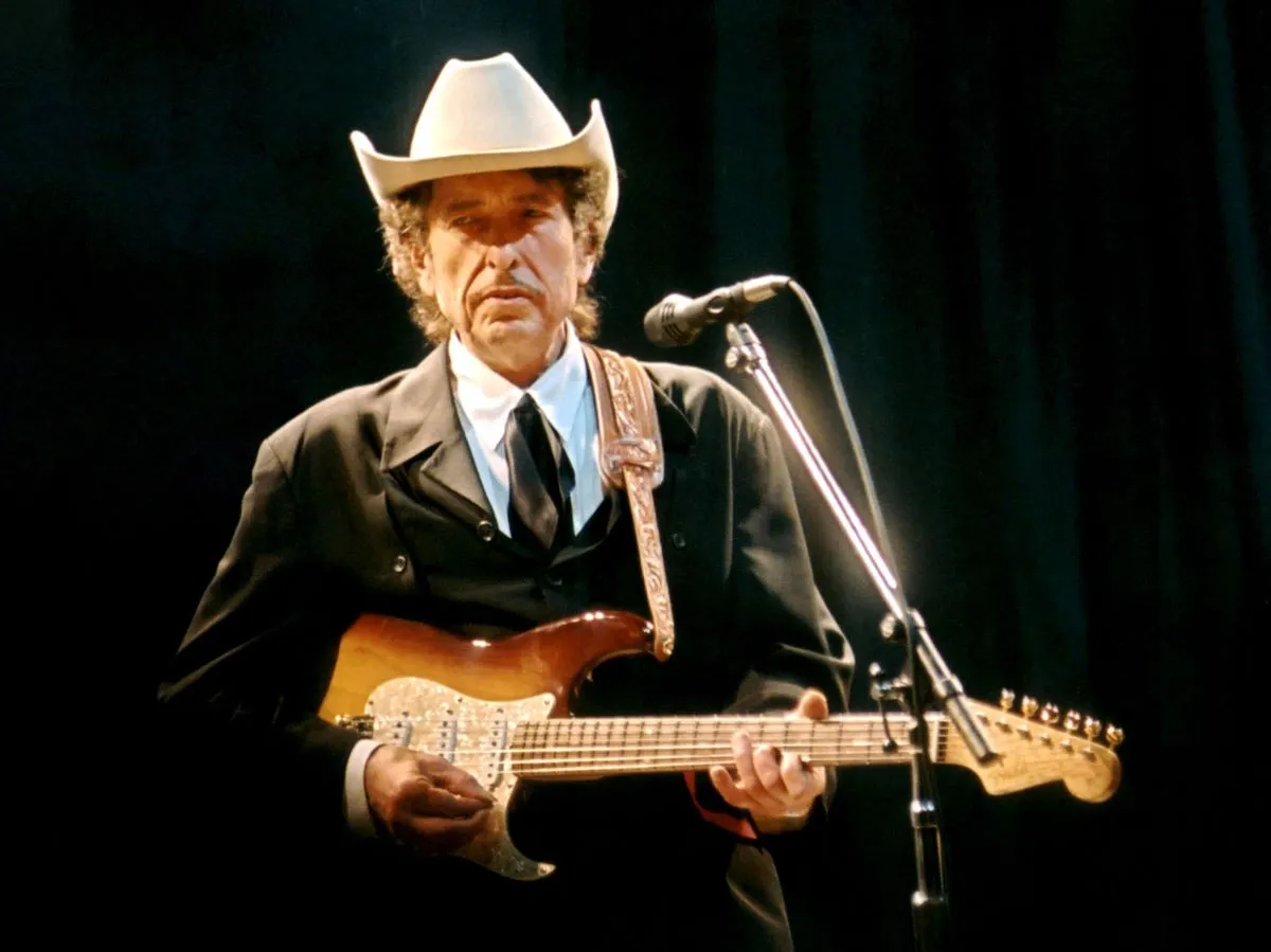 Bob Dylan wears a suit and a cowboy hat. He holds an electric guitar and stands in front of a microphone.