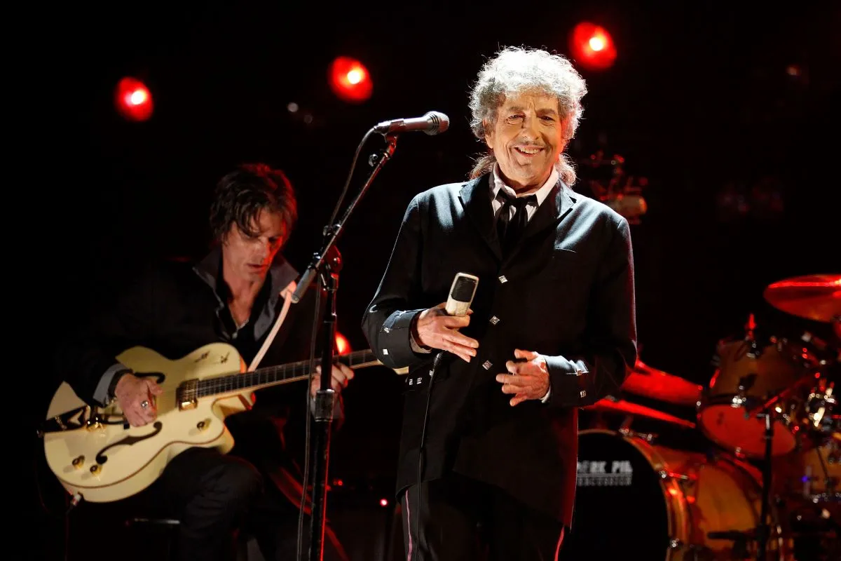 Bob Dylan holds a microphone and stands on a stage.