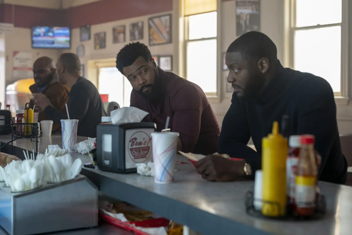 Isaiah Mustafa as John Sampson and Aldis Hodge as Alex Cross sitting at the counter at Ben's Chili Bowl in Prime Video's 'Cross'