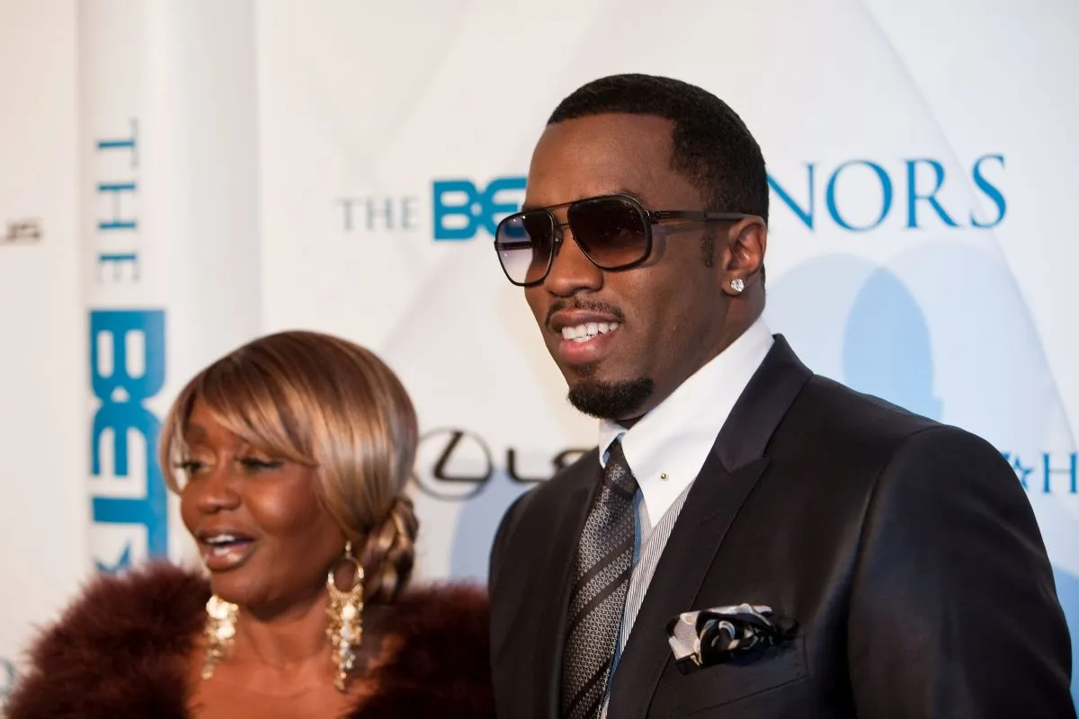 Sean 'Diddy' Combs wears a suit and sunglasses. He stands with his mother, who wears a fur coat and gold earrings.