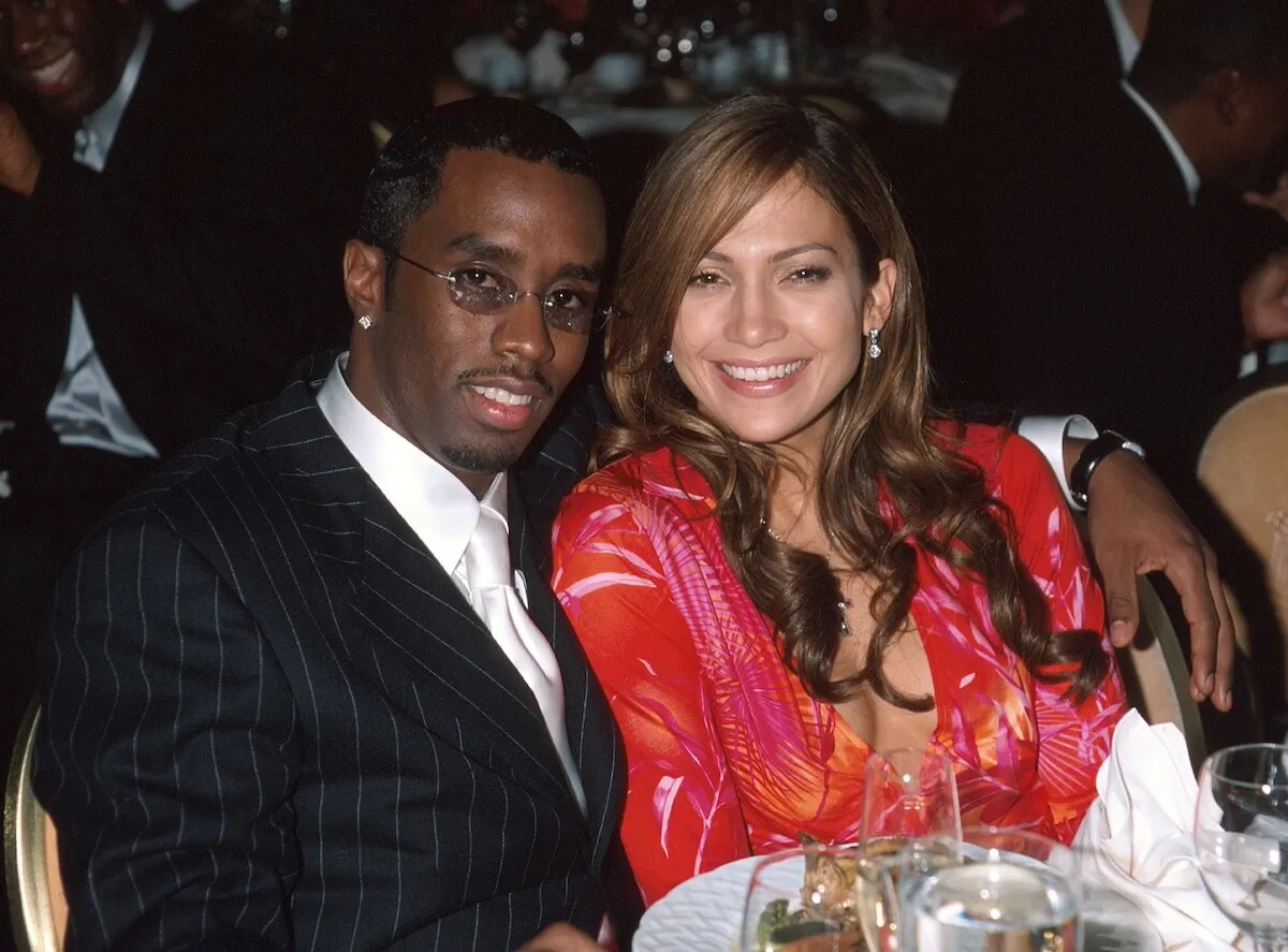 Sean 'Diddy' Combs and Jennifer Lopez sit at a table together. He wears a suit and she wears a pink shirt.