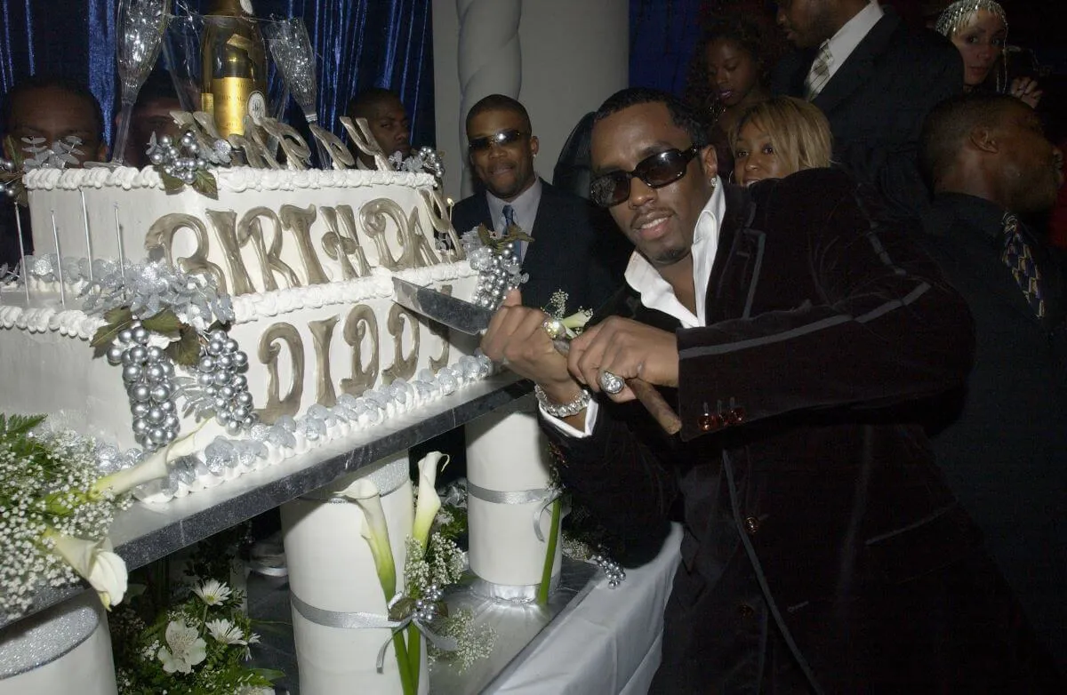 Sean "Diddy" Combs wears sunglasses and a suit. He holds a knife in front of a cake that says "Happy Birthday Diddy" in gold letters.