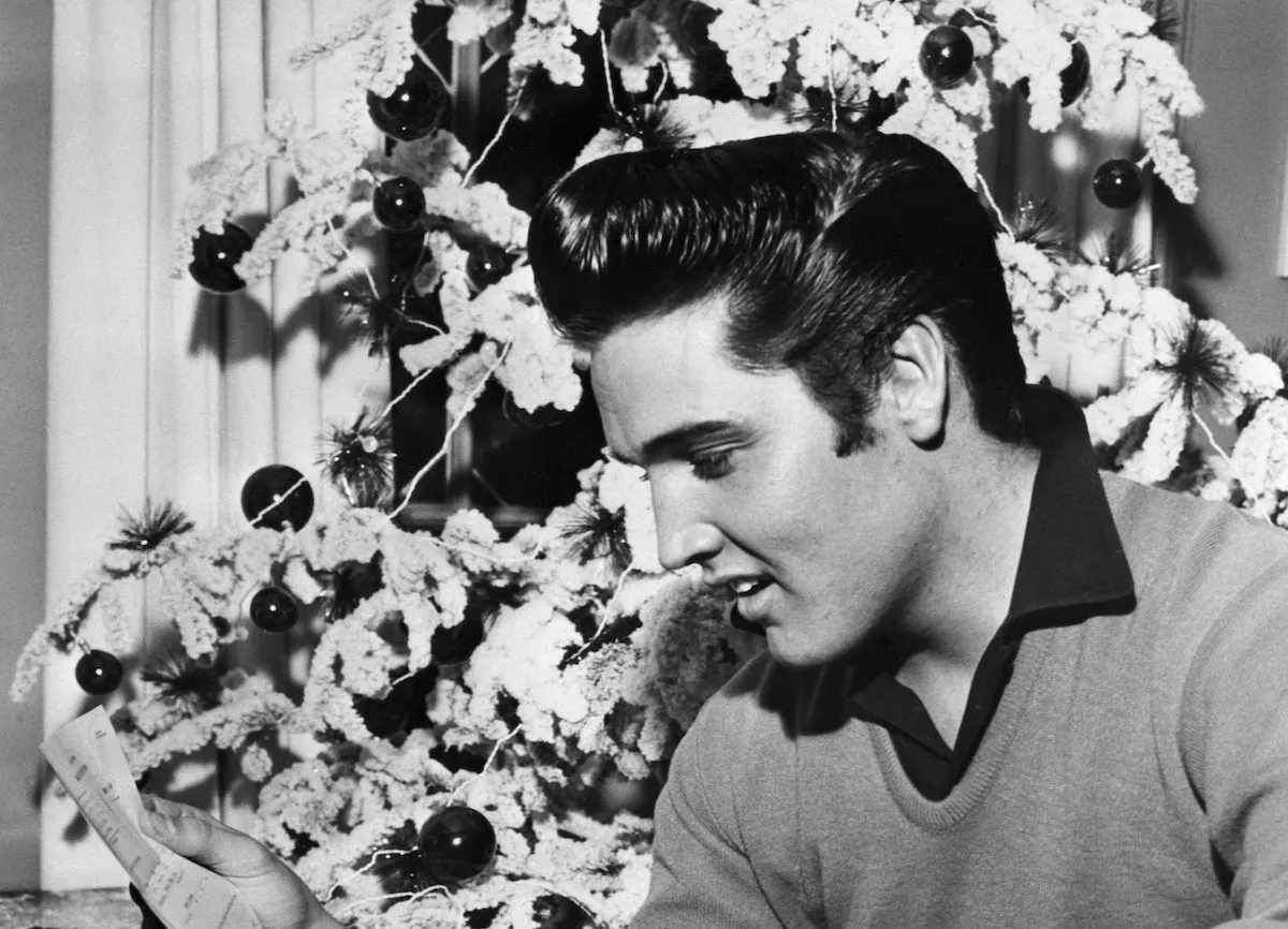 Black and white photo of a young Elvis sitting in front of a flocked Christmas tree and reading a letter