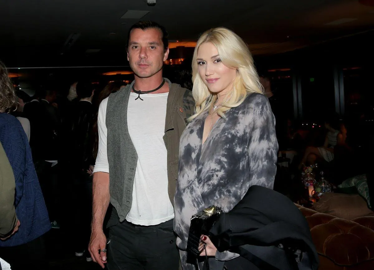 Gavin Rossdale and Gwen Stefani stand together. He wears a white shirt with a green vest and she wears a gray tie-dye shirt.