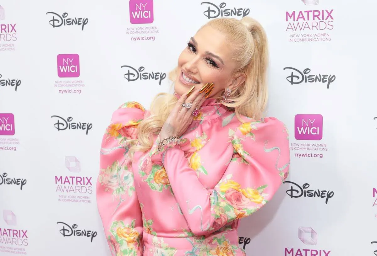 Gwen Stefani wears a pink dress and stands in front of a white background. She touches her hand to her face.