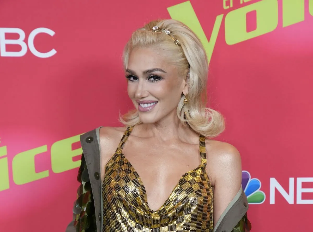 Gwen Stefani wears a checkered dress and stands in front of signs for 'The Voice.'