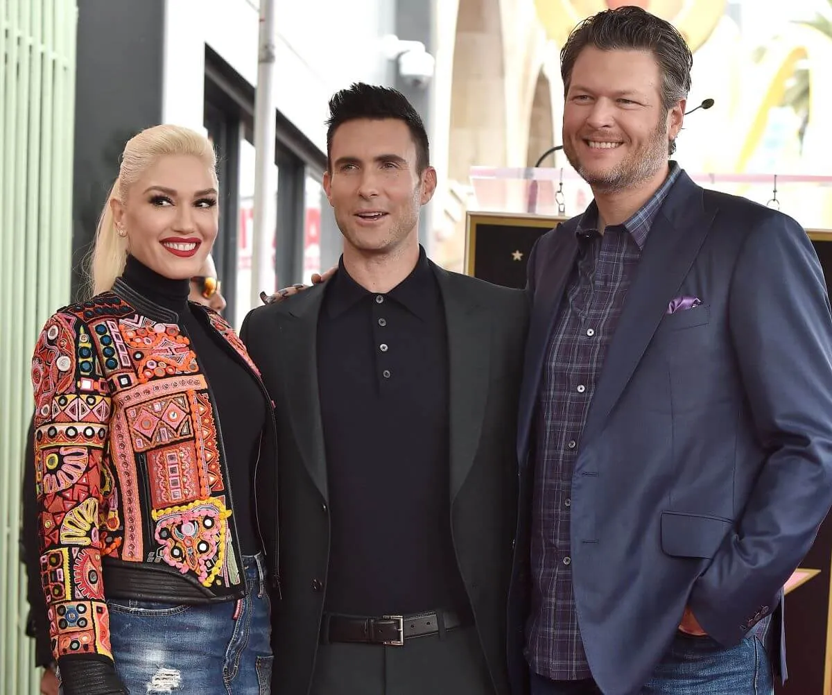 Gwen Stefani, Adam Levine, and Blake Shelton stand together. Stefani wears a patterned jacket, Levine wears a black suit, and Shelton wears a blue suit.