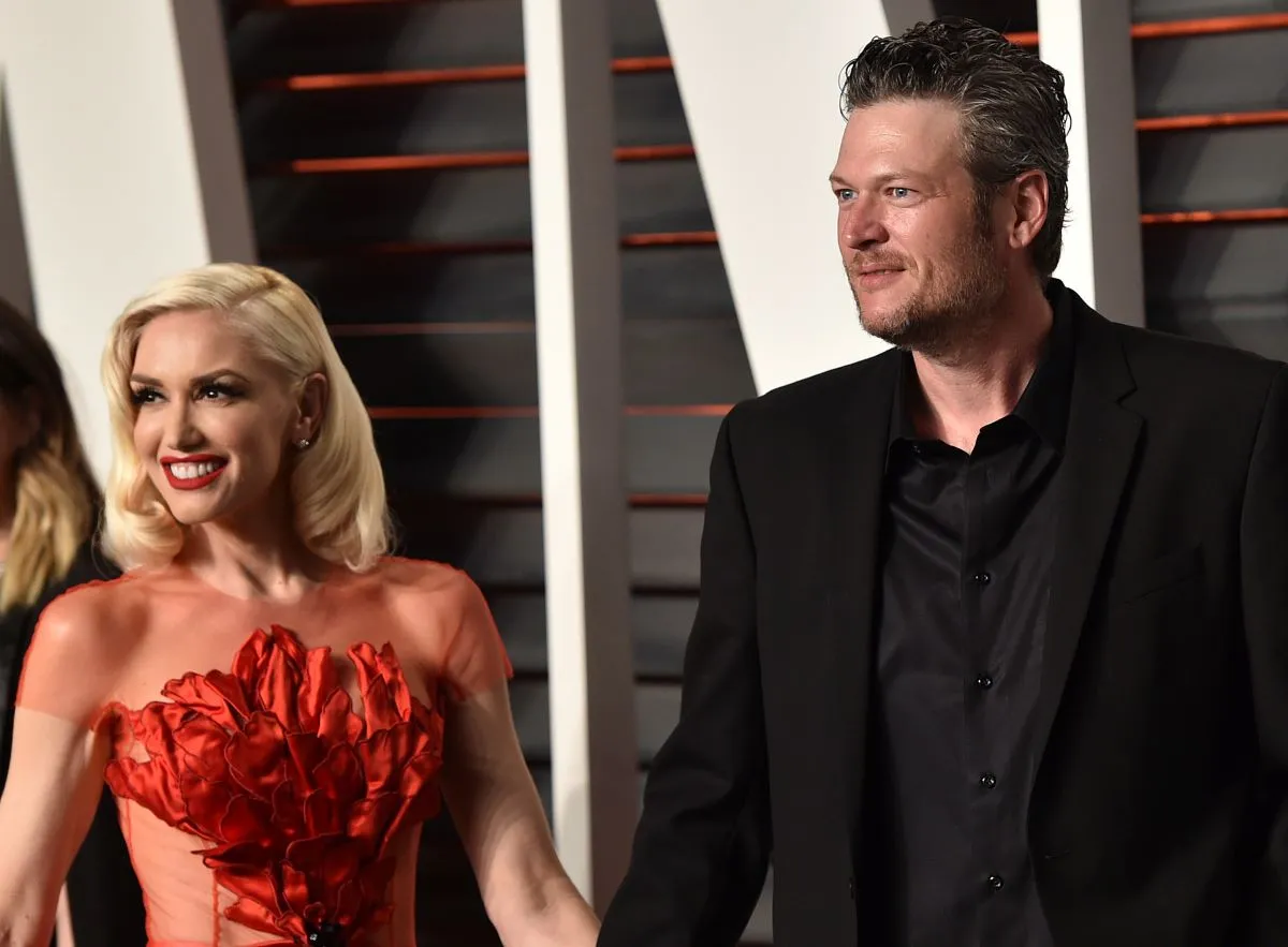 Gwen Stefani wears a red dress and holds Blake Shelton's hand. He wears a black suit.