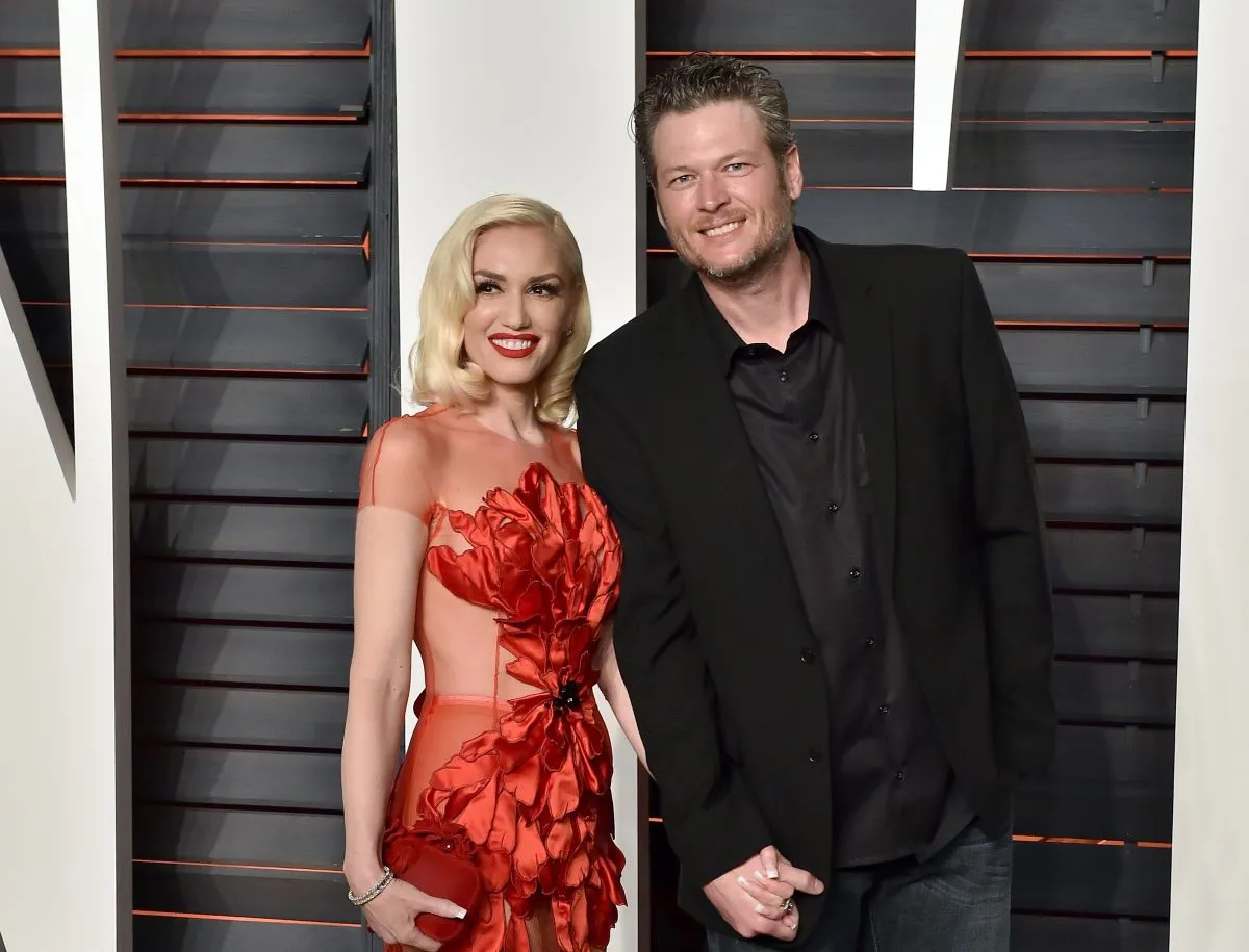 Gwen Stefani wears a red dress and holds Blake Shelton's hand. He wears a black suit.