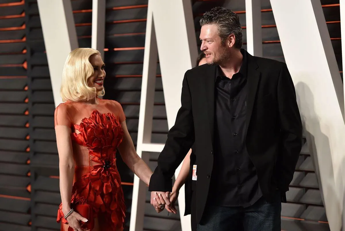 Gwen Stefani and Blake Shelton hold hands. She wears a red dress and he wears a black suit.