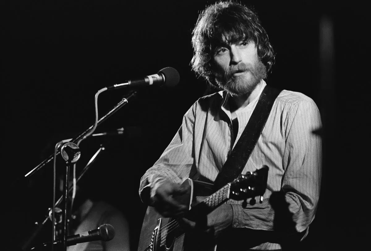 A black and white picture of JD Souther playing guitar and standing in front of a microphone.