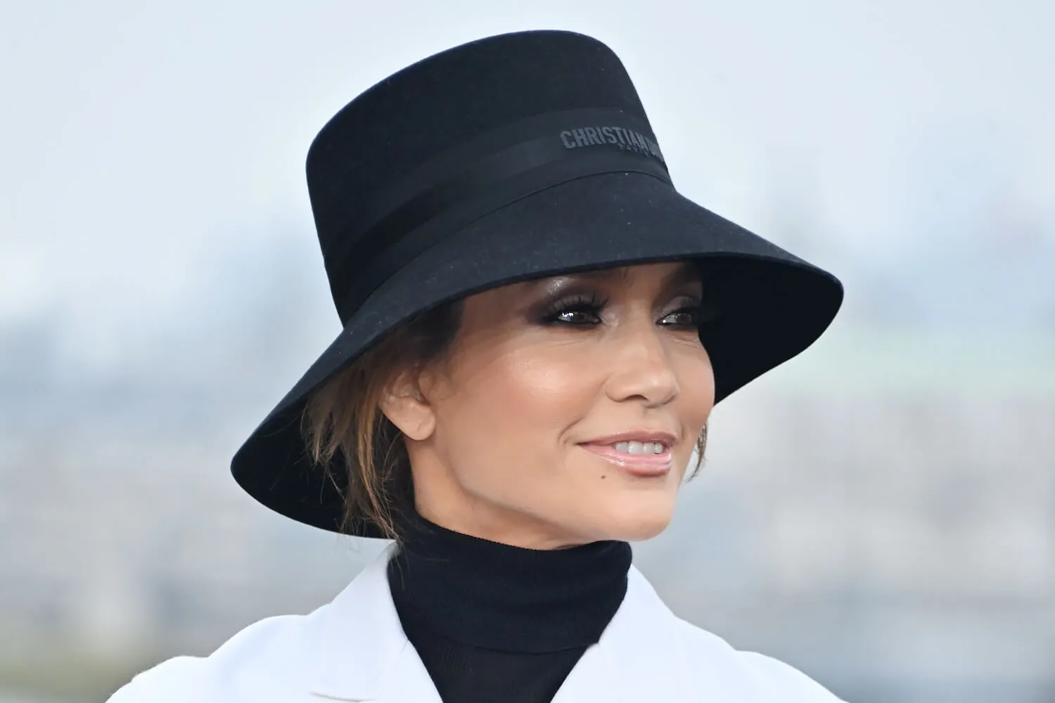 Jennifer Lopez attends the London photocall for 'Unstoppable.' She's looking over her left shoulder and wearing a white coat, a black shirt underneath, and a black brimmed hat.