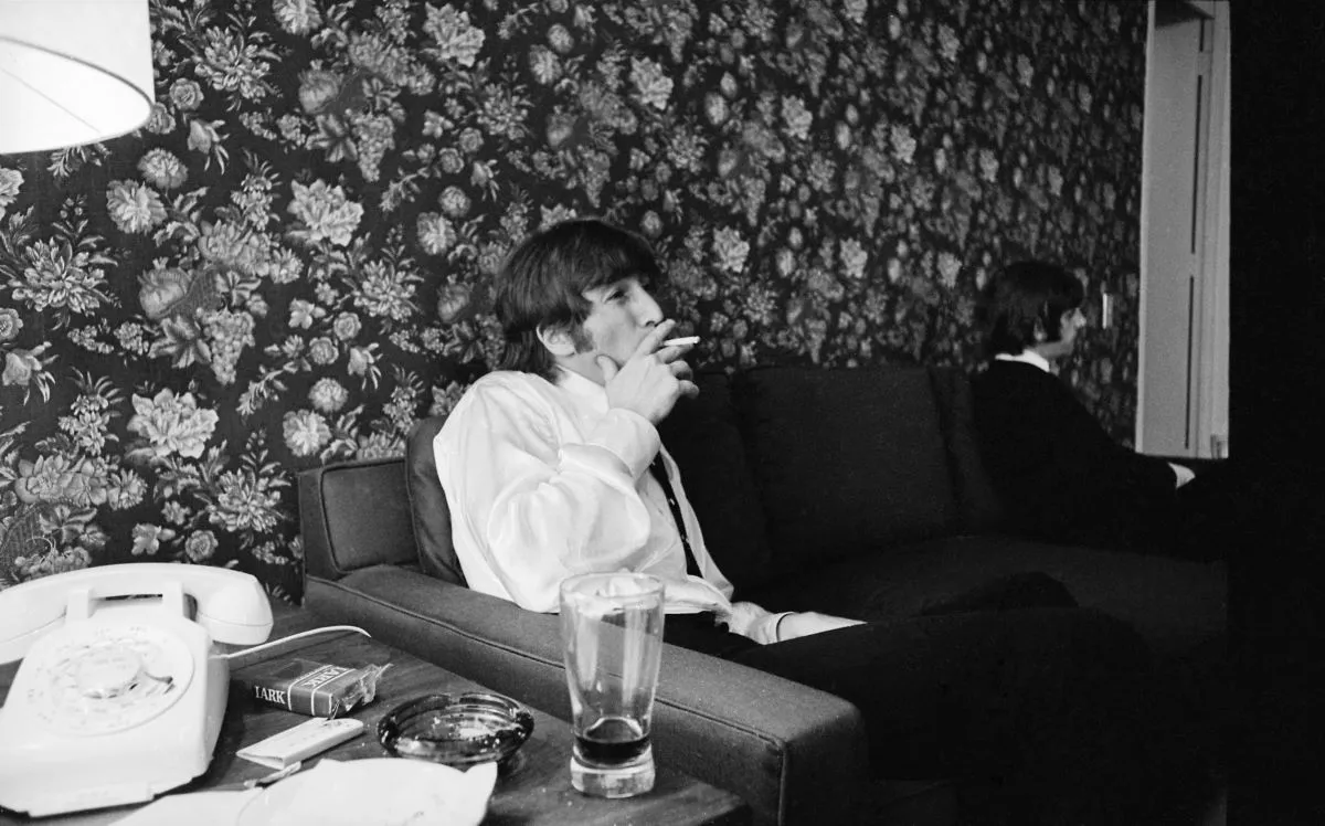 John Lennon sits on a couch and smokes a cigarette.