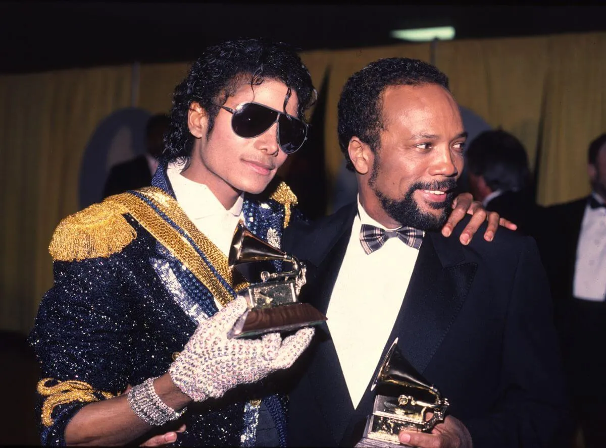 Michael Jackson wears a sparkling jacket, gloves, and sunglasses. He stands with his hand on Quincy Jones' shoulder. They both hold Grammys.