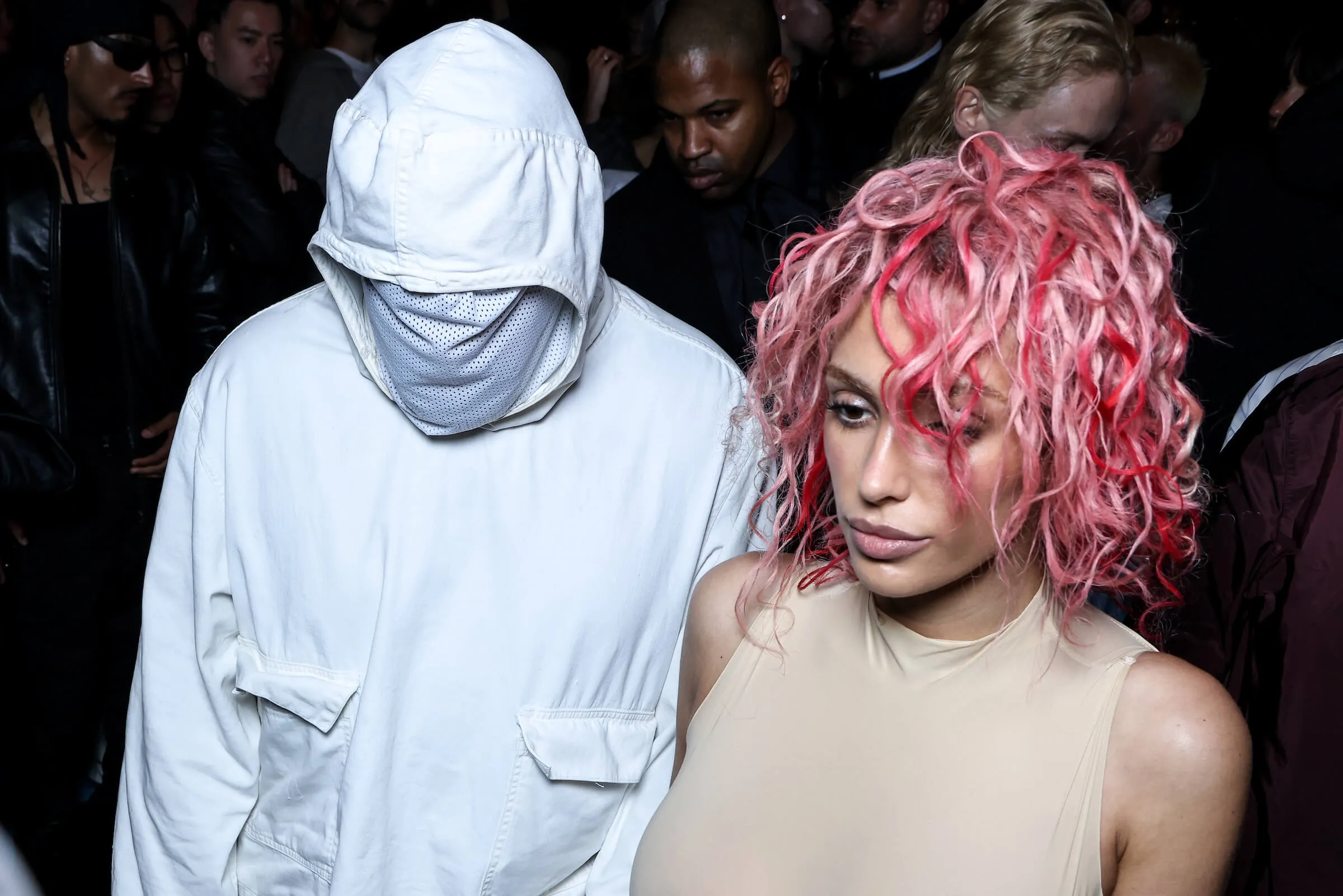 Kanye West and Bianca Censori walking together at a fashion show. West is wearing head-to-toe white with a white face covering. Censori is wearing a nude bodysuit and pink hair.