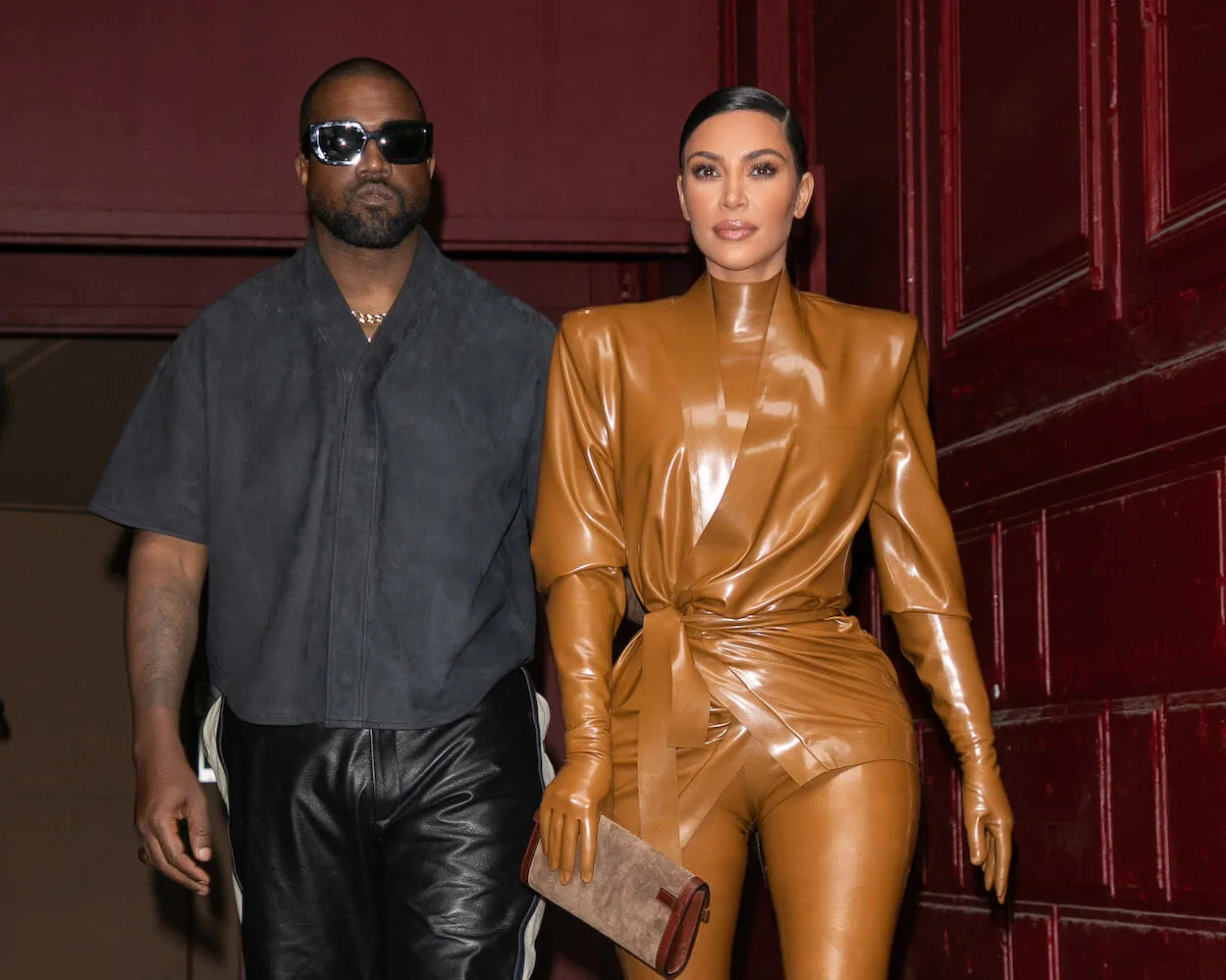 Kanye West and Kim Kardashian walking together in 2020 at Paris Fashion Week. West is wearing a short-sleeve black shirt with black pants. Kardashian is wearing a gold bodysuit with gloves.