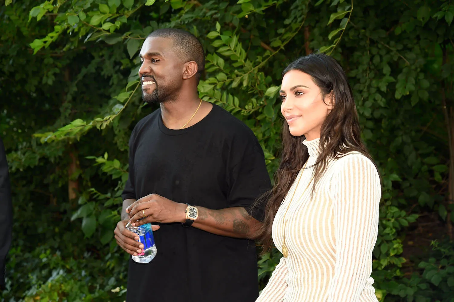 Kanye West and Kim Kardashian standing next to each outside. West is smiling and holding a water bottle and Kardashian is smiling with her mouthed closed.