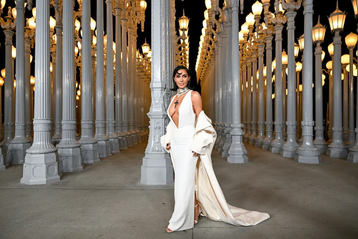 Kim Kardashian at the 2024 LACMA Art+Film Gala at the Los Angeles County Museum of Art. She's posing in a white sleeveless, low-cut dress with a large cross necklace and a cream coat that's fallen to her elbows.