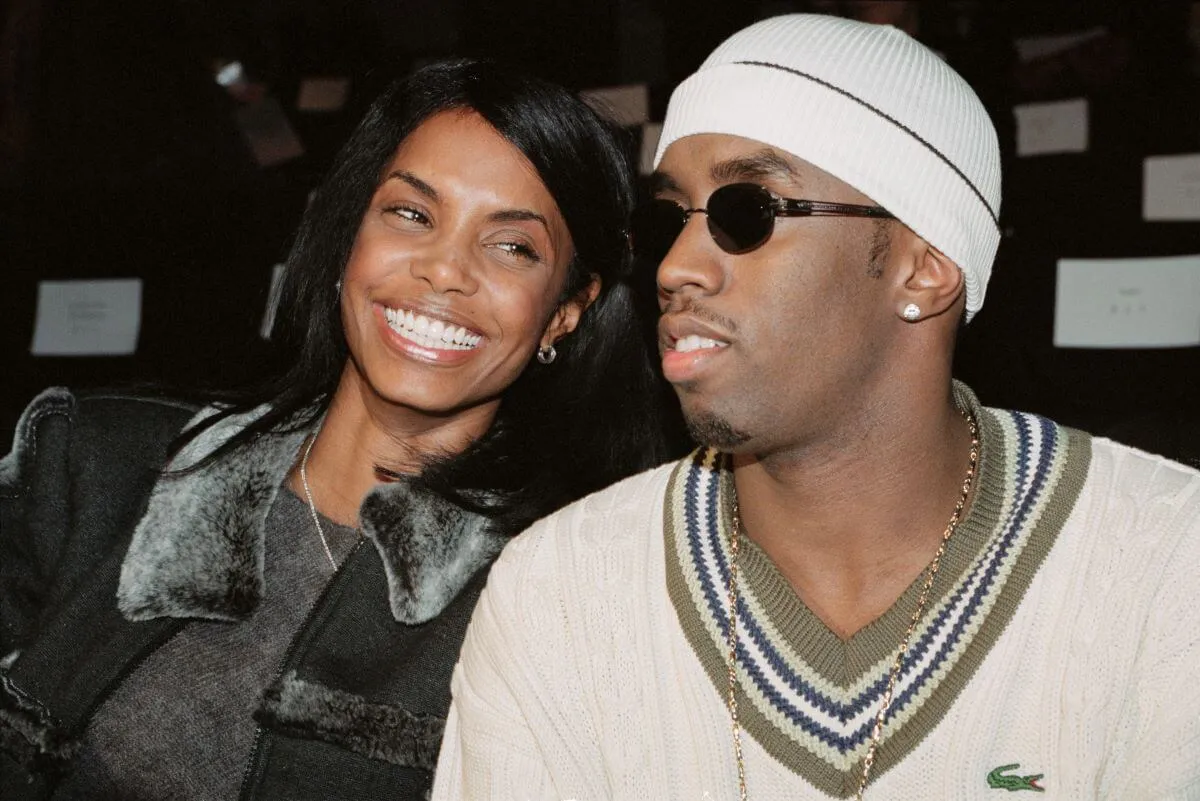 Kim Porter and Sean 'Diddy' Combs sit together. She wears a jacket and smiles. He wears a white sweater, hat, and sunglasses.