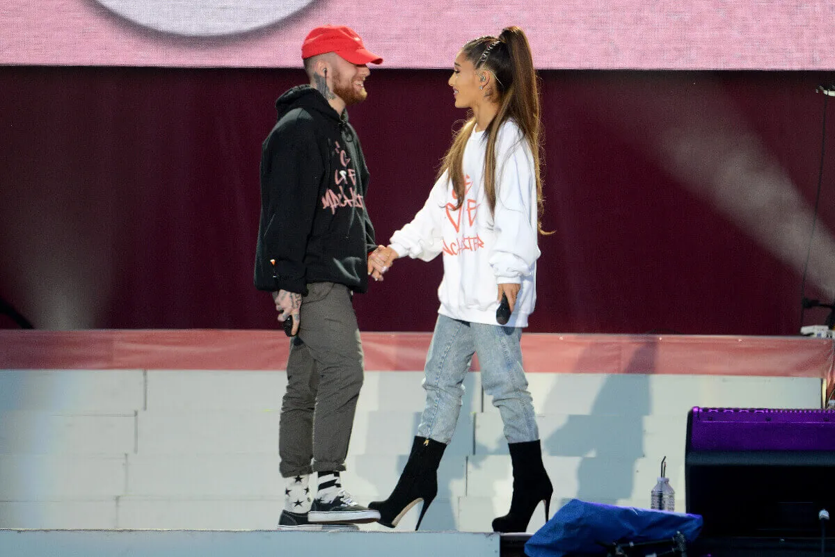 Mac Miller and Ariana Grande stand on a stage together. He wears an orange hat and black sweatshirt and she wears a white sweatshirt.