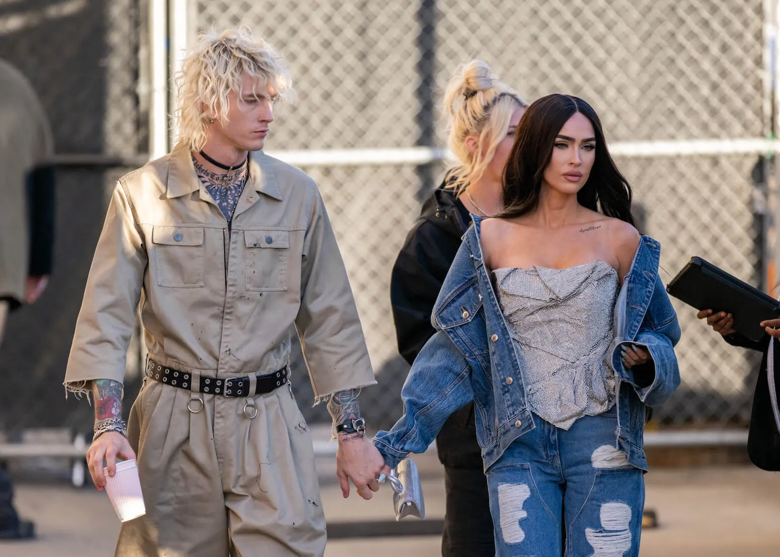 Machine Gun Kelly and Megan Fox walking in Los Angeles while holding hands. Fox is wearing a denim jacket and jeans, and Kelly is wearing a khaki coverall.