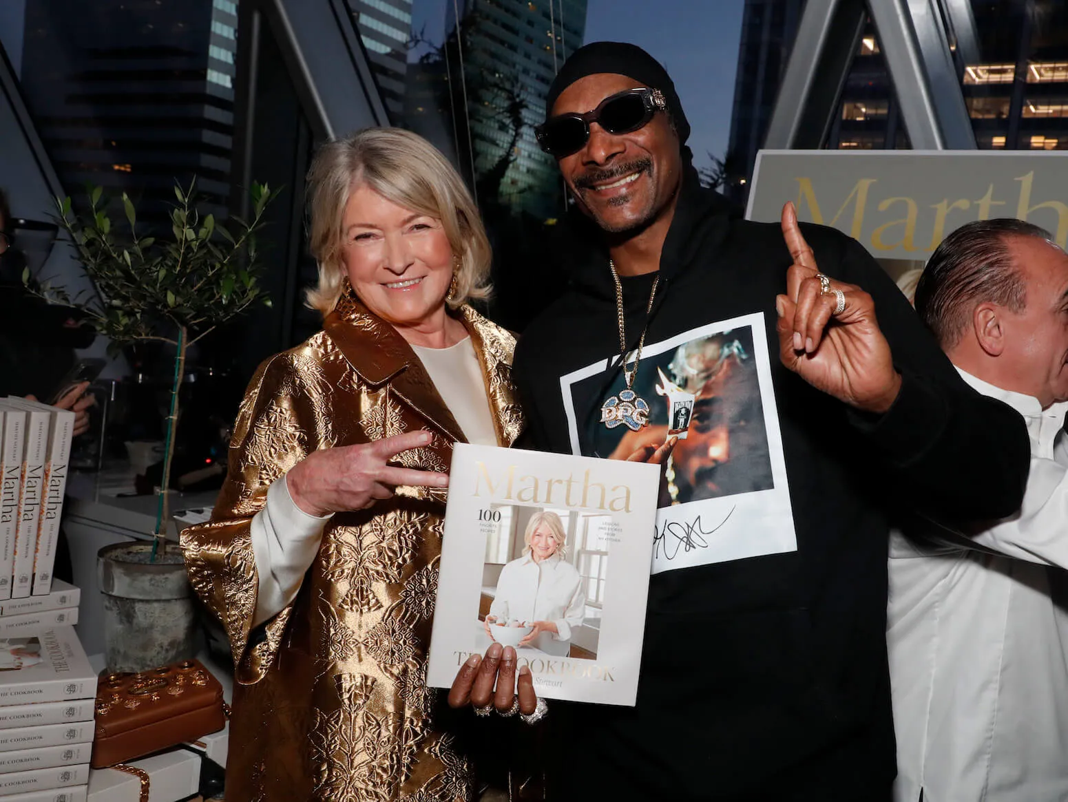 Martha Stewart holding her book and smiling while next to Snoop Dogg, who's also smiling. Stewart has her hand in a peace sign; Snoop has his pointer finger up.