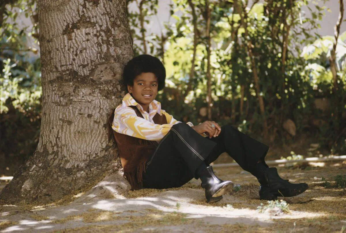 A young Michael Jackson sits under a tree.