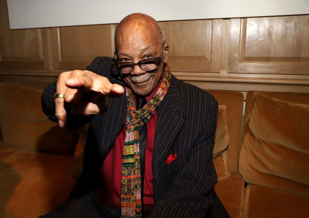 Quincy Jones wears a suit and a scarf. He sits on a brown couch and points at the camera.