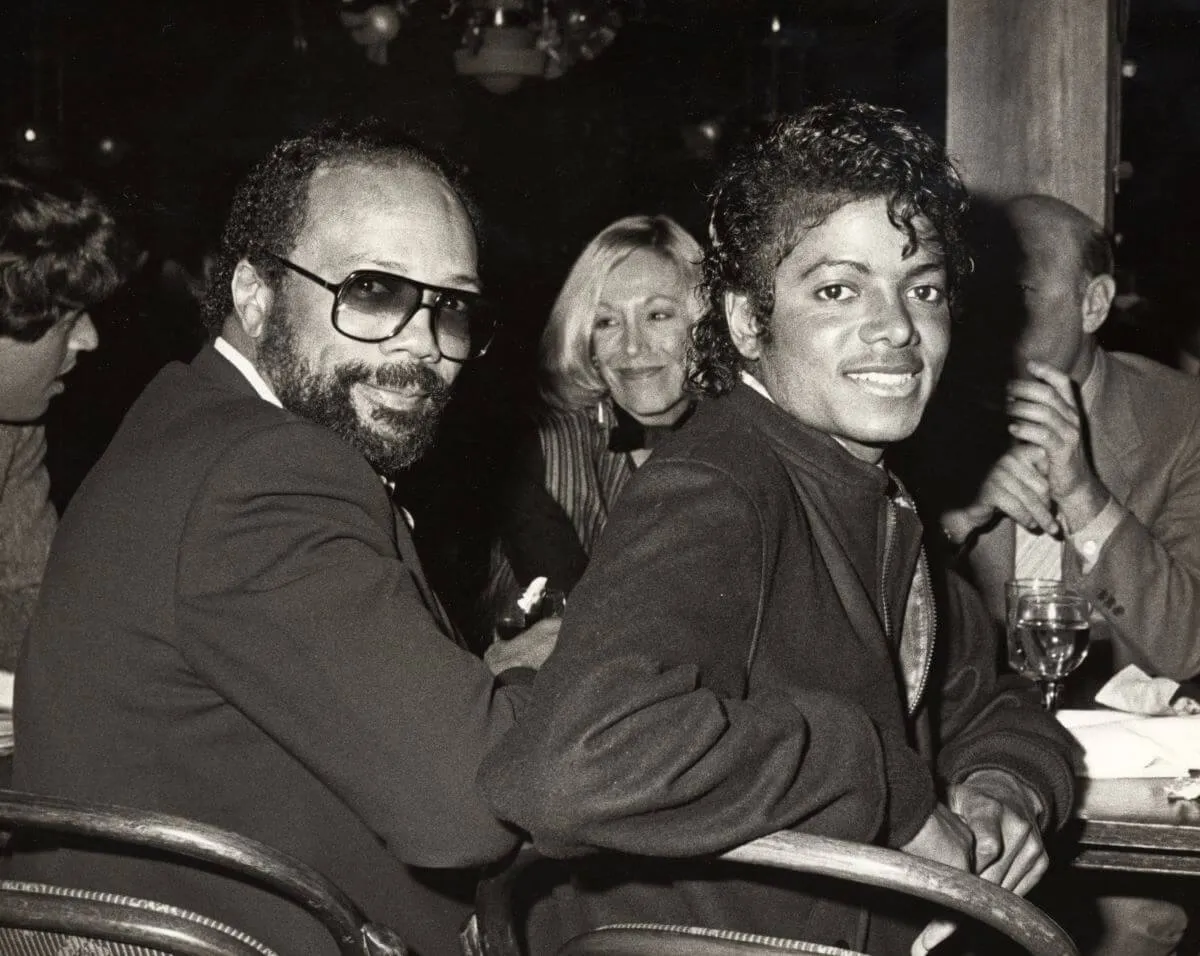 A black and white picture of Quincy Jones and Michael Jackson sitting at a table. They turn to face the camera.