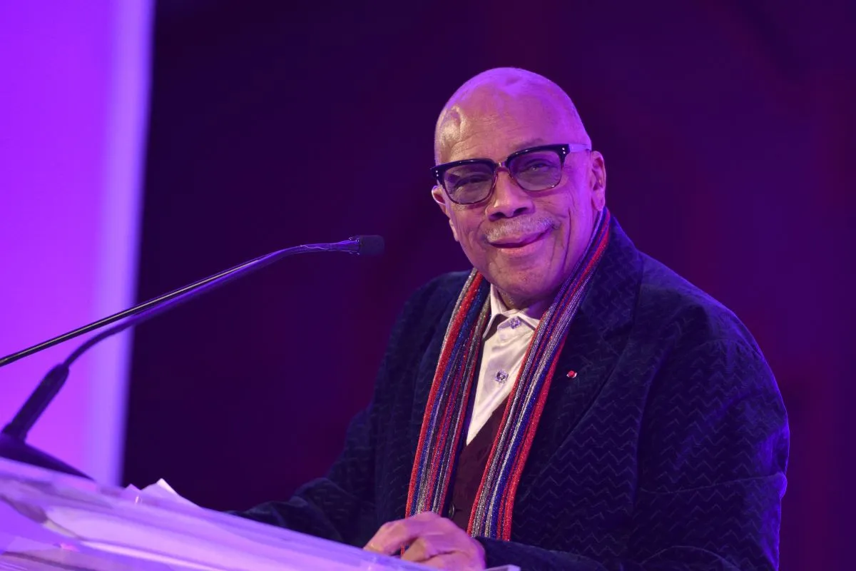 Quincy Jones wears a suit and a colorful scarf. He stands at a podium.