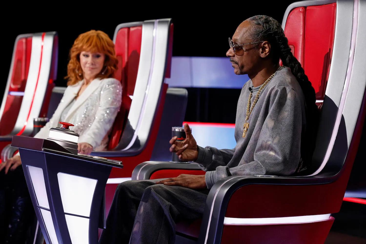 Reba McEntire out of focus sitting in a red chair on 'The Voice' while Snoop Dogg is in the foreground and in focus in a red chair. 