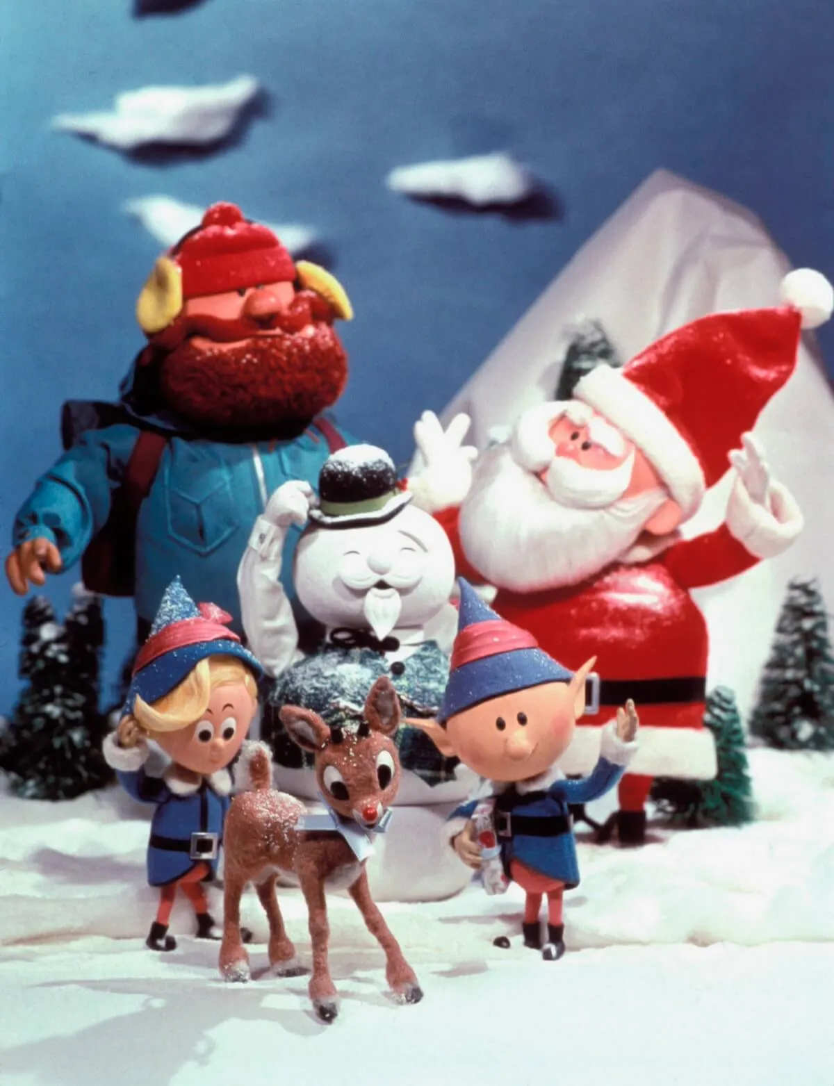 Image of stop-motion characters from 'Rudolph The Red-Nosed Reindeer' standing in a group, including Santa, Rudolph, Yukon Cornelius, the Snowman, and elves