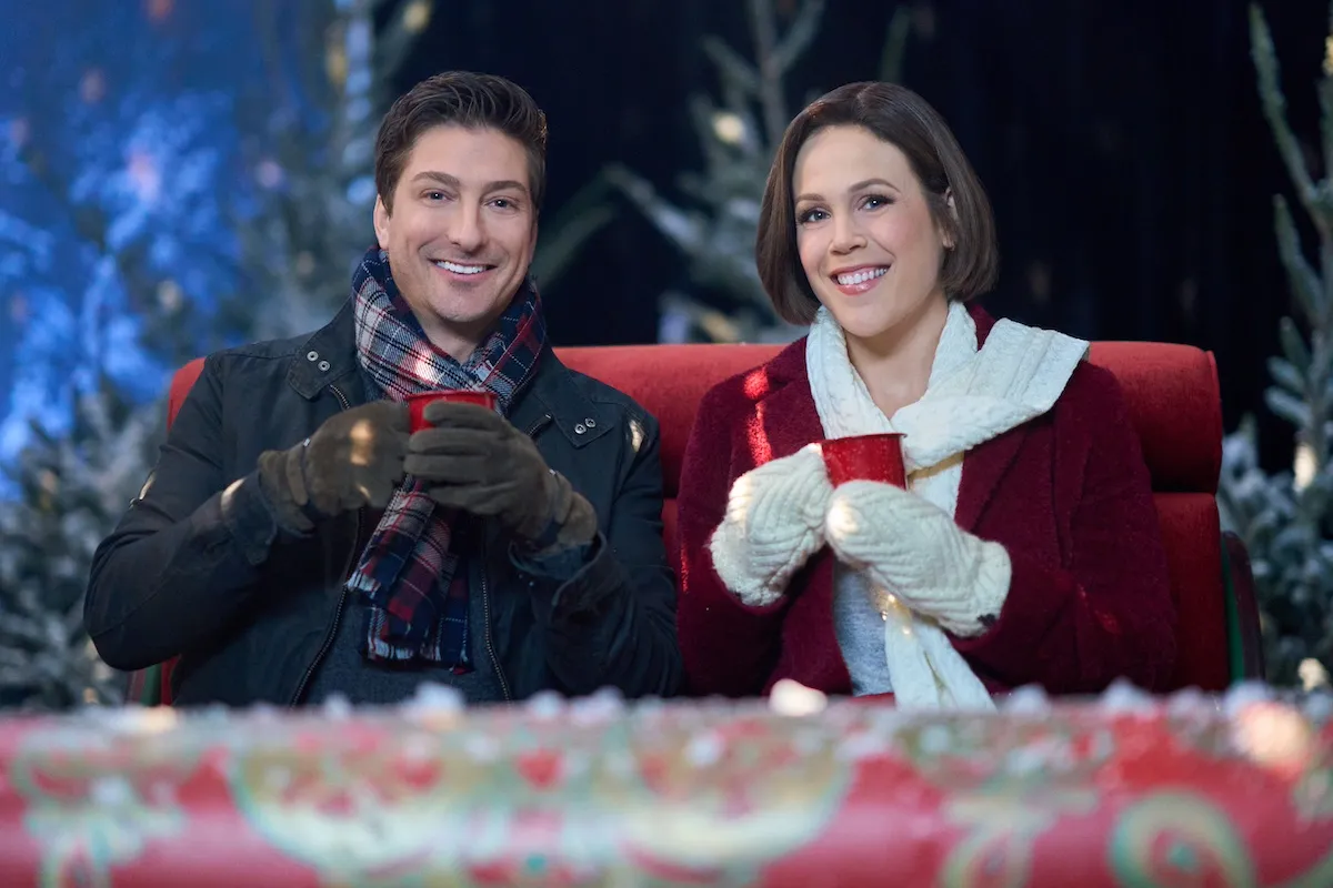 Erin Krakow, wearing a red jacket and white scarf and holding a red mug, sitting next to Daniel Lissing, also holding a red mug, in the Hallmark movie 'Santa Tell Me'