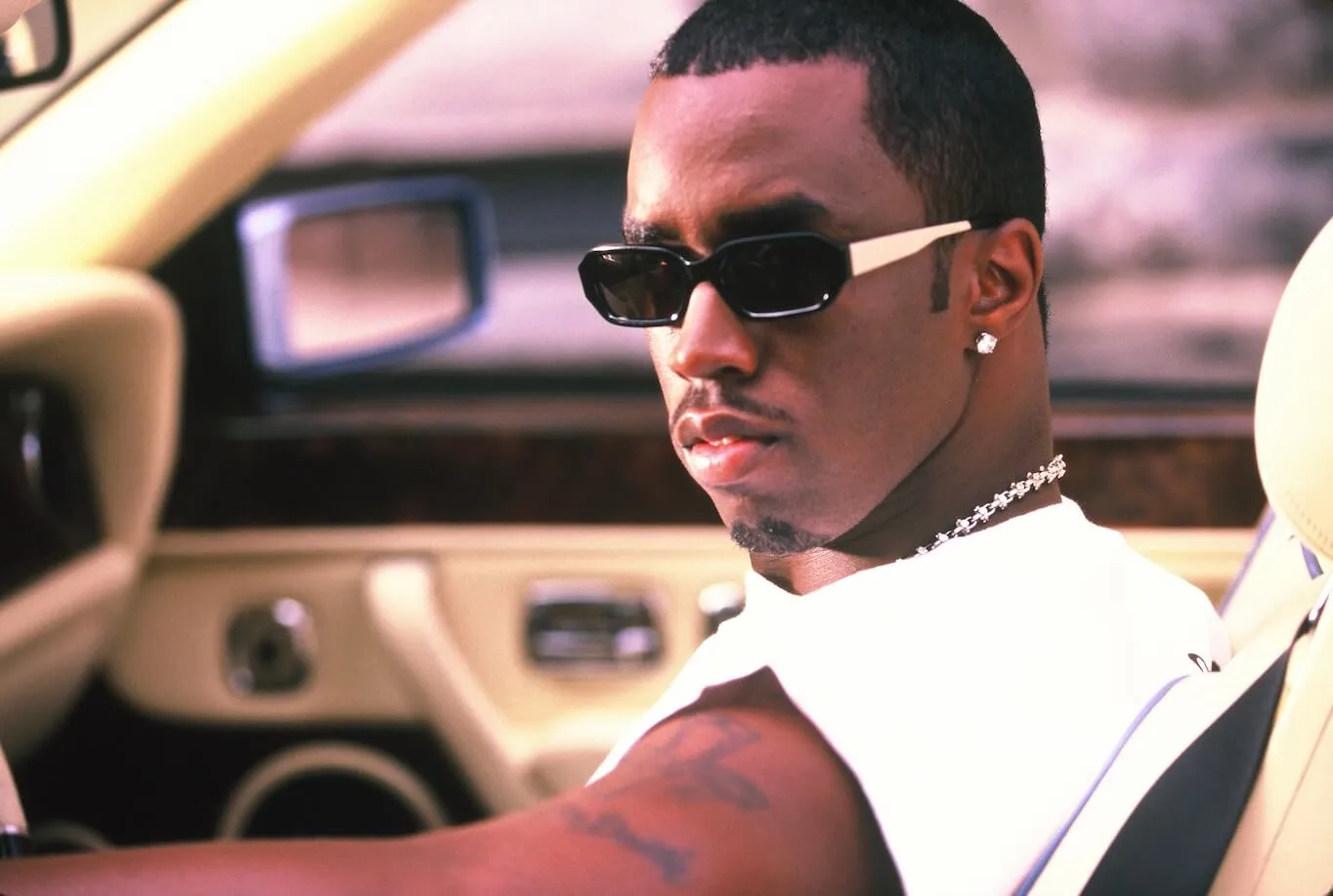 A close-up of Sean 'Diddy' Combs looking over his left shoulder while driving a car in a white tank top and sunglasses in 1998.