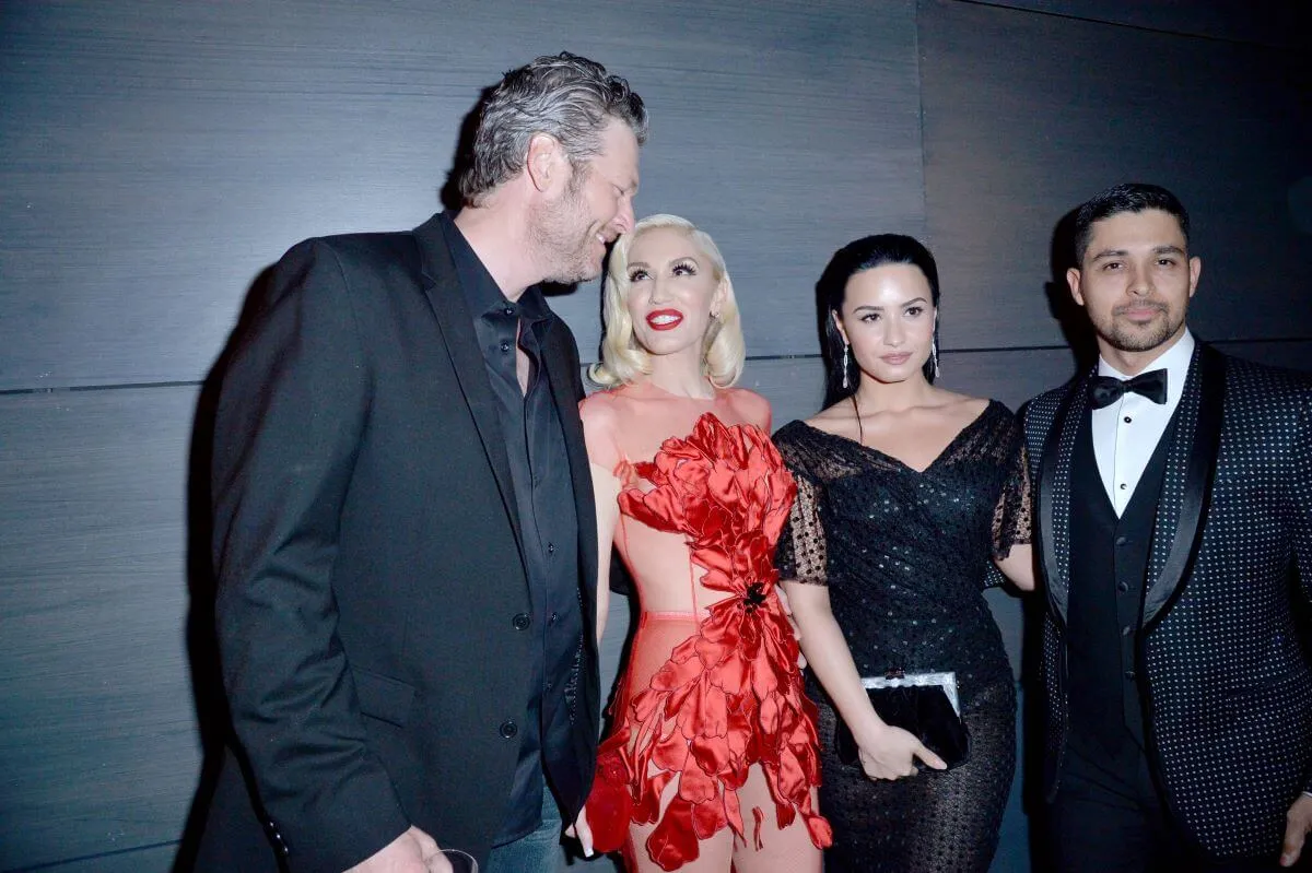 Blake Shelton, Gwen Stefani, Demi Lovato, and Wilmer Valderrama stand together. Shelton, Lovato, and Valderrama wear black and Stefani wears red.