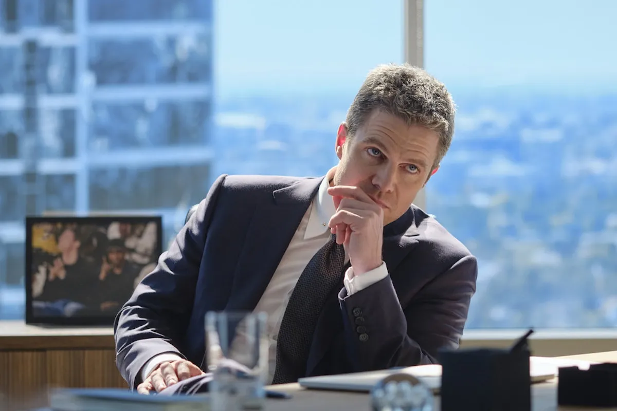 Stephen Amell sitting at a desk with his chin in his hand and the city skyline in the windows behind him in the 'Suits LA' pilot