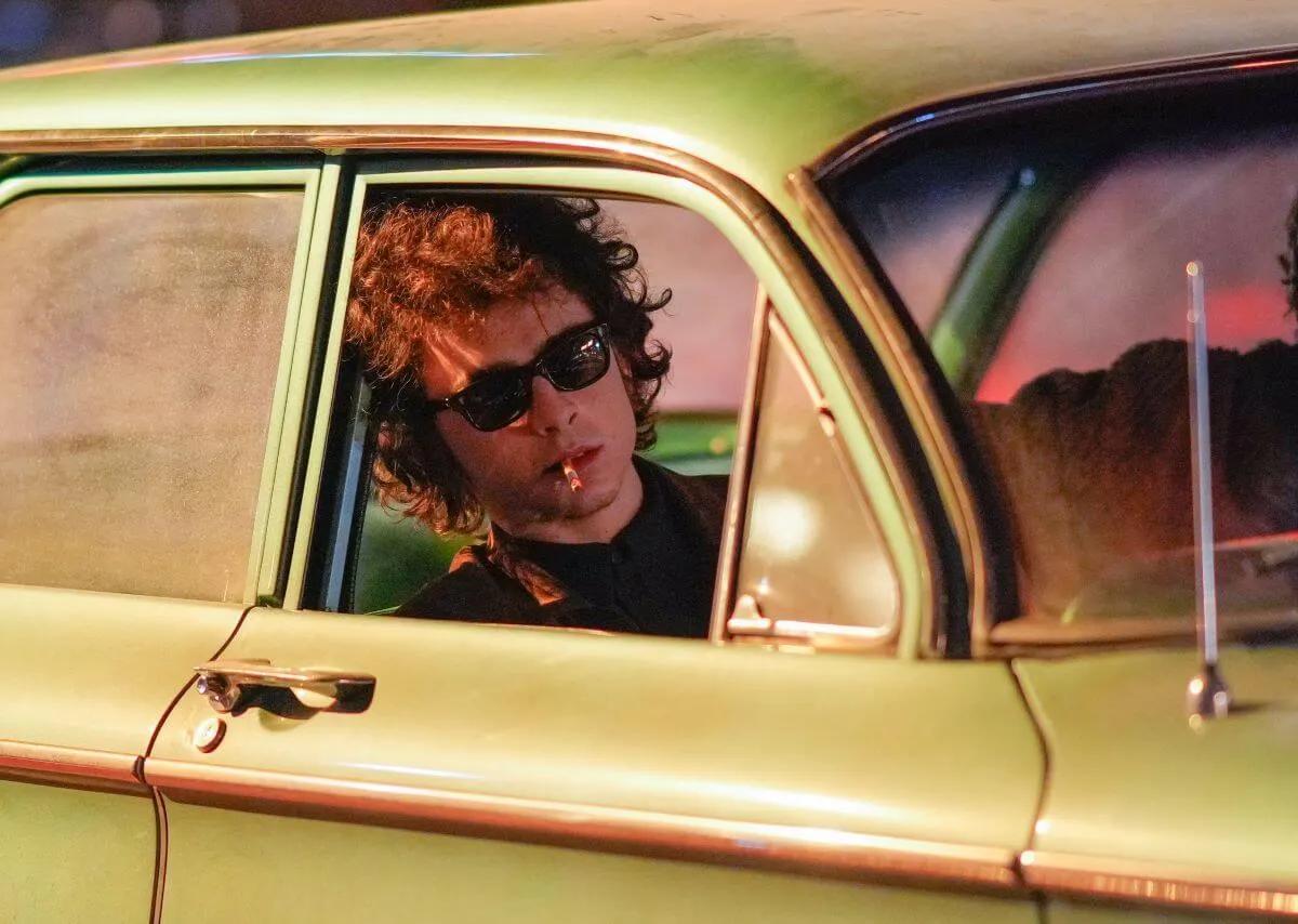 Timothee Chalamet wears sunglasses and sits in a car. He has a cigarette in his mouth.