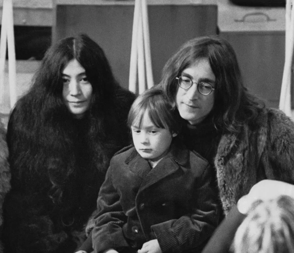 A black and white picture  of Yoko Ono, Julian Lennon, and John Lennon sitting together.