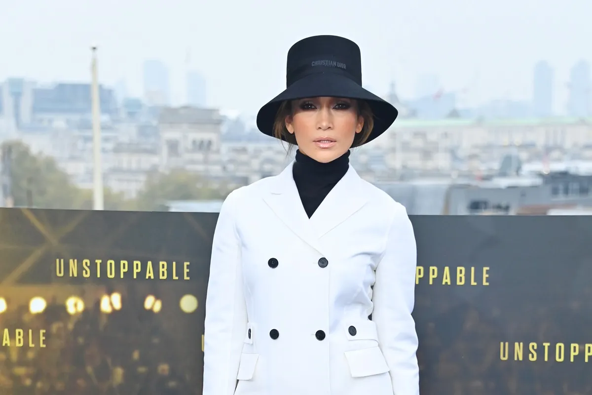 Jennifer Lopez posing at the premiere of 'Unstoppable' in a white trench coat and black hat.