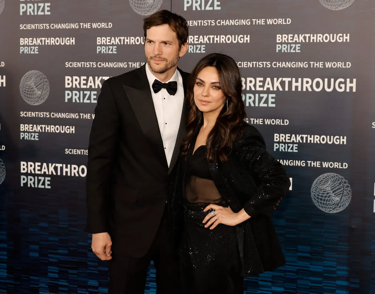 Ashton Kutcher and Mila Kunis stand together. He wears a tuxedo and she wears a black dress.